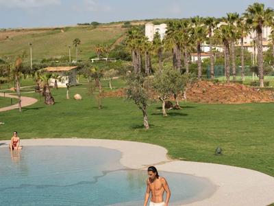 Žena na okraji lagunového bazénu s zeleným trávníkem a palmami v pozadí.