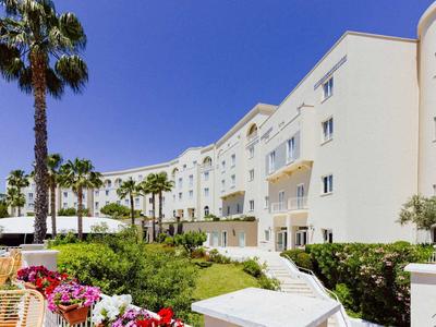 Moderní bílá hotelová budova s palmami a kvetoucími květinami pod jasně modrou oblohou.