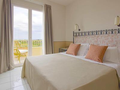 Světý hotelový pokoj s velkou postelí, hnědými polštáři a výhledem na balkon.