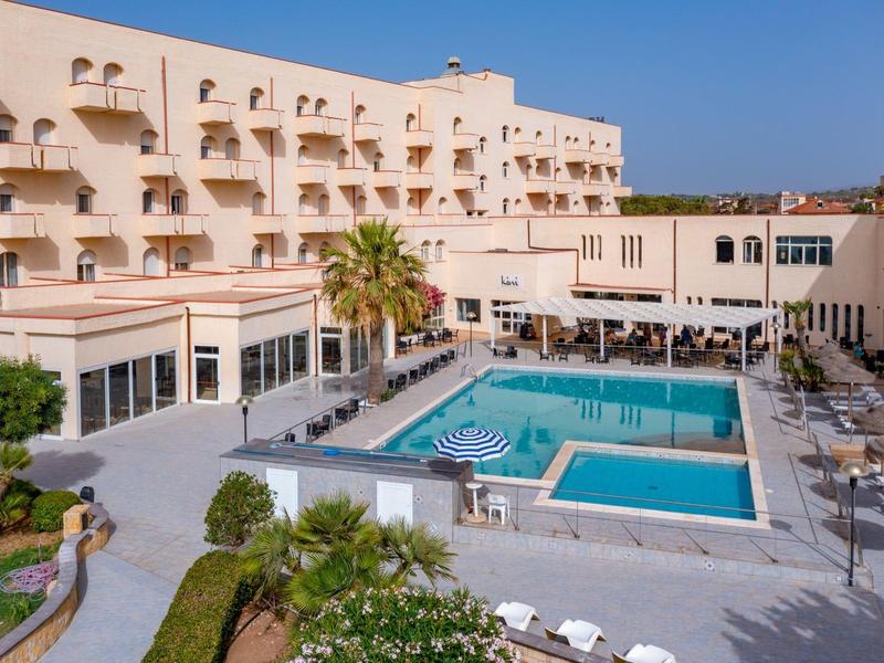 Hotel with large outdoor pool and lounge chairs under clear blue sky.