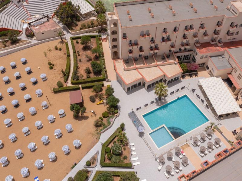 Aerial view of a beach resort with sandy beach, umbrellas, and a rectangular pool next to the hotel.