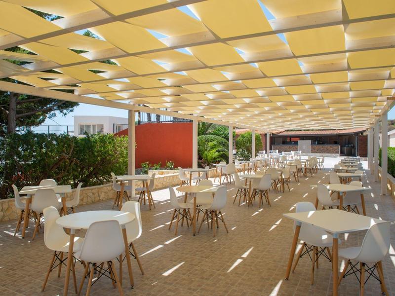 Large covered outdoor area with many white tables and chairs, surrounded by plants.