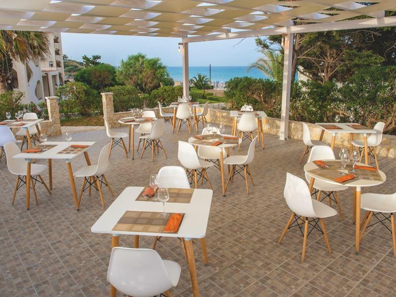 Outdoor dining area with white chairs and tables, sea and palm trees in the background.