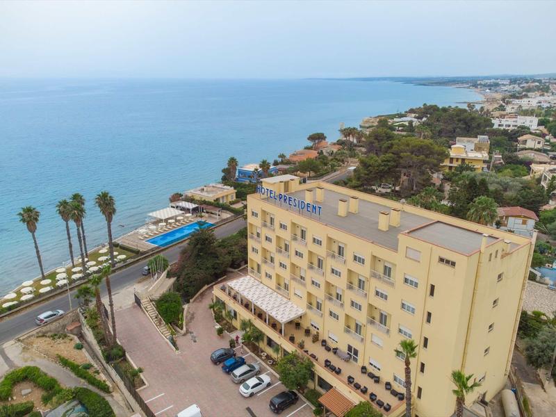 Widok na żółty hotel przy wybrzeżu z basenem, parkingiem i palmami nad morzem.