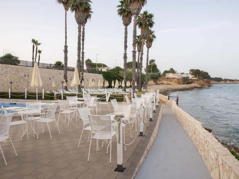 Puste białe krzesła i stoliki na tarasie z widokiem na morze i palmy.