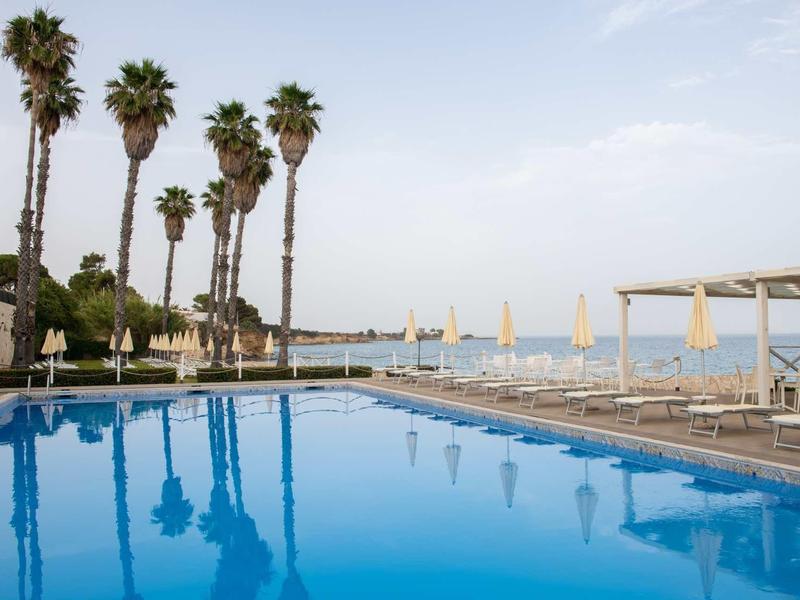 Niebieski basen z palmami i leżakami z widokiem na morze pod niebieskim niebem.