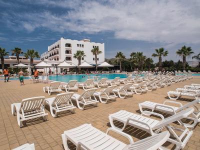 Viele weiße Sonnenliegen am Pool eines Hotels mit Palmen und blauem Himmel.