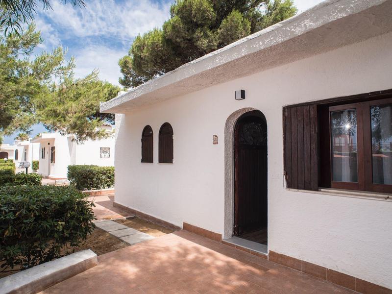 Weißes Ferienhaus mit Fensterläden und Bäumen bei klarem Himmel.