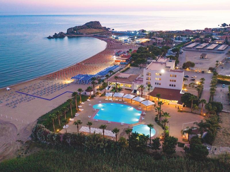 Blick auf einen beleuchteten Pool und Strand bei Dämmerung am Meer mit Gebäuden und Hügeln im Hintergrund.
