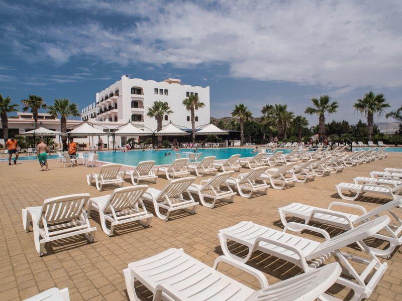Viele weiße Sonnenliegen am Pool eines Hotels mit Palmen und blauem Himmel.