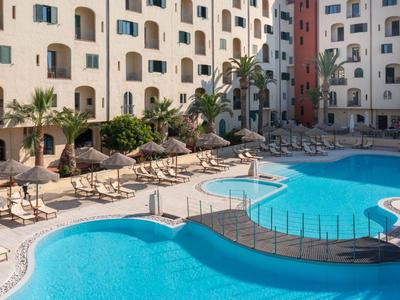 Groot hotelzwembad met ligstoelen en parasols naast een meerlaag gebouw.