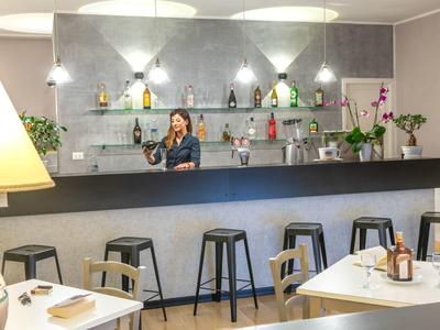 Moderne Bar mit dunkelgrauem Tresen, schwarzen Hockern, hellen Wänden und dekorativen Pflanzen.