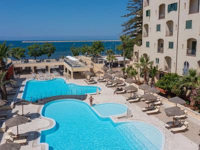 Großer blauer Hotelpool mit Liegestühlen und Palmen, dahinter Meer und blauer Himmel.