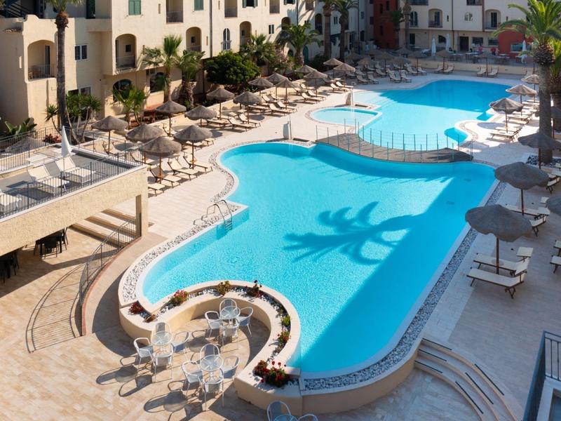 Klares blaues Schwimmbecken mit Liegestühlen, Sonnenschirmen und umliegenden Gebäuden bei Sonnenschein.