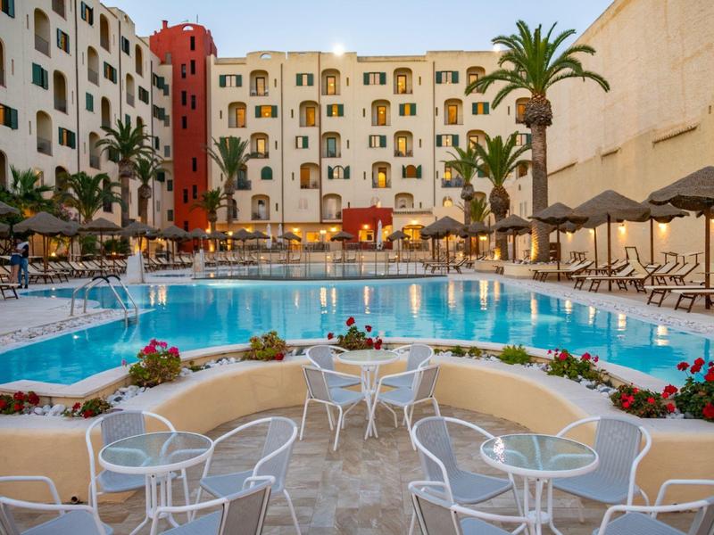 Großer Pool mit blauer Wasserfläche, umgeben von weißen Stühlen, Sonnenschirmen und Palmen vor Hotel.