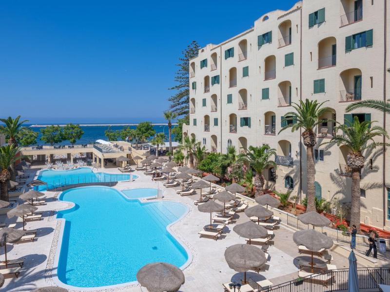 Großer Außenpool mit Sonnenliegen neben mehrstöckigem Hotel und Meer im Hintergrund bei klarem Himmel.