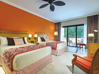 Hotelzimmer mit zwei Doppelbetten, orangener Wand und Balkon mit Aussicht auf grüne Bäume.