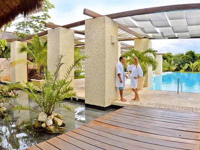 Moderner Außenbereich mit Holzweg, Wasserbecken, Säulen, Pool und grüner Palmenumgebung unter klarem Himmel.