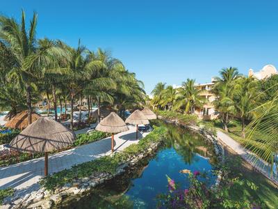 Tropischer Hotelgarten mit Palmen, Sonnenschirmen und Wasserlauf bei klarem Himmel.