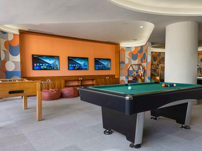 Moderner Spielbereich mit Billardtisch, Tischfußball und drei Bildschirmen an orangefarbener Wand.