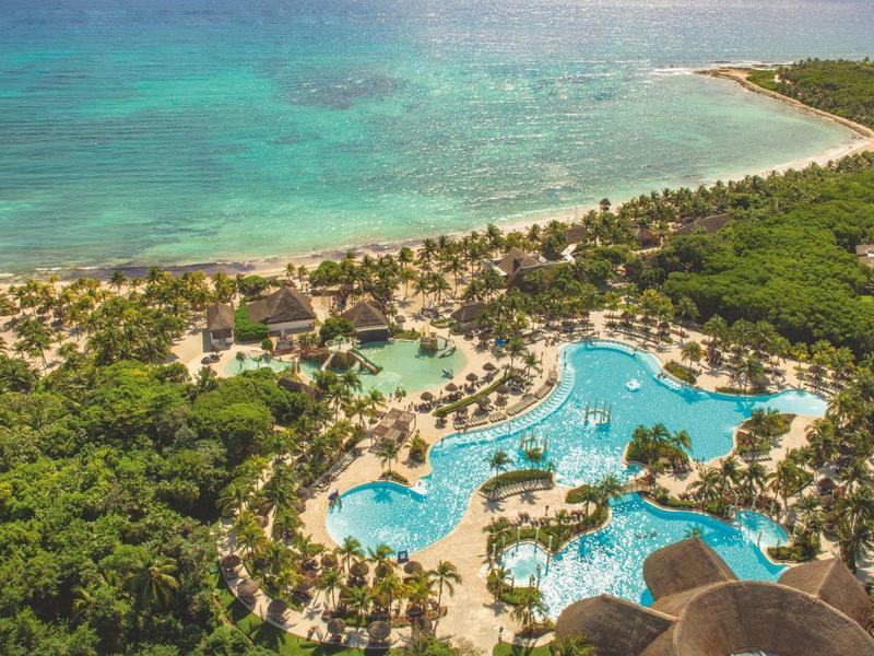 Hotelanlage mit großen Pools neben einem tropischen Strand und türkisfarbenem Meer.