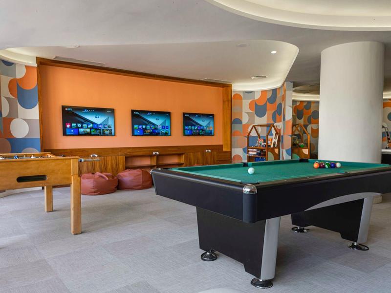 Moderner Spielbereich mit Billardtisch, Tischfußball und drei Bildschirmen an orangefarbener Wand.
