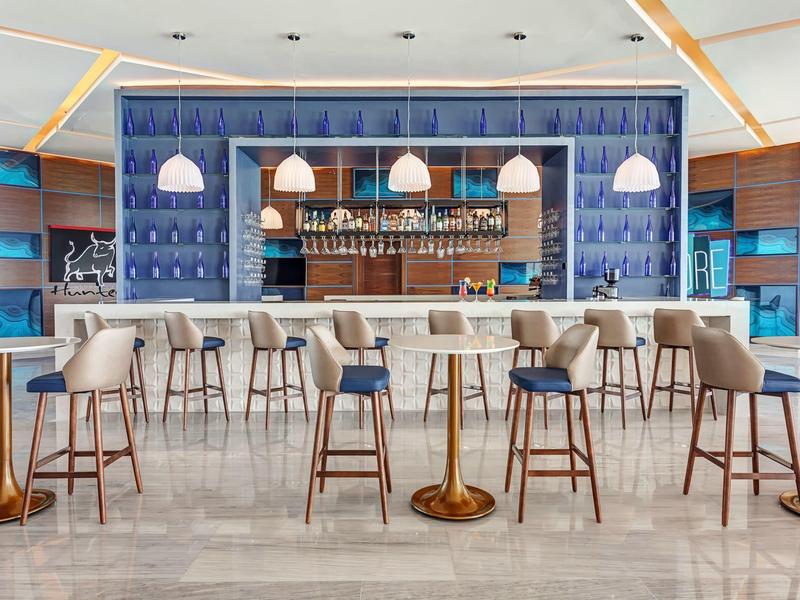 Moderner Hotelbarbereich mit blauen Wänden, hölzernen Stühlen und Pendelleuchten über der Bar.