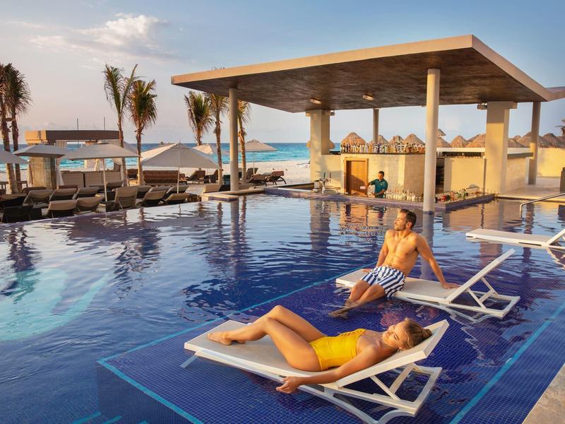 Freizeitpool mit blauer Kachel, Liegen, Palmen und Meerblick bei Sonnenuntergang.