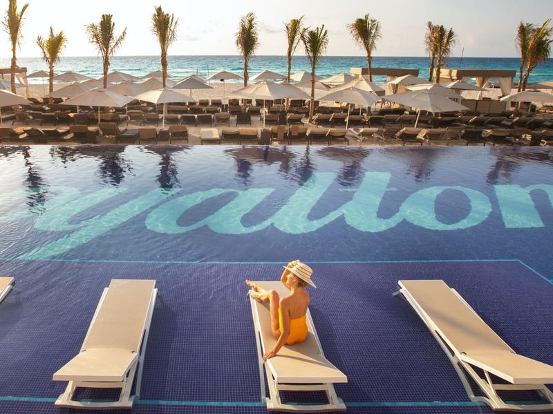 Blick auf Pool mit Schriftzug, Sonnenliegen, Palmen und Meer im Hintergrund bei Sonnenuntergang.