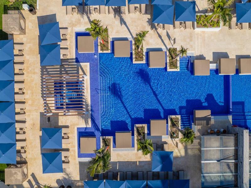 Luftaufnahme von großem blauem Pool mit weißen Liegestühlen und grünen Pflanzen am Rand.