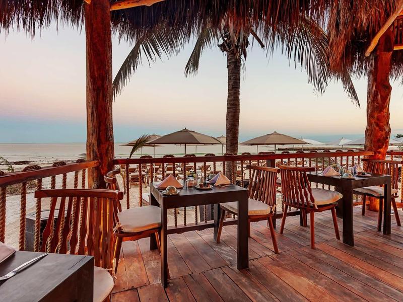 Holzterrasse mit Stühlen und Tischen, Blick auf Meer und Strand, Palmenblätter als Dach.