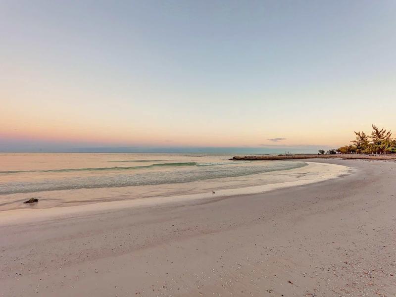Breiter Sandstrand mit sanften Wellen, weiter Horizont und pastellfarbener Himmel bei Sonnenuntergang.