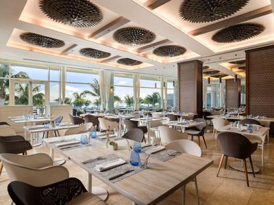 Modernes Hotelrestaurant mit Holztischen, schwarzen Stühlen und großen Fenstern mit Blick auf Palmen.