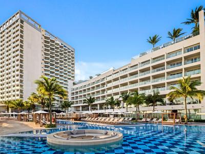 Modernes Hotel mit großen Pools, Sonnenschirmen und Palmen bei klarem Himmel.