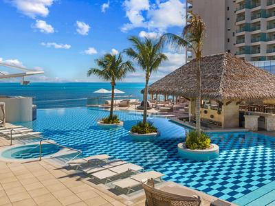 Hotelpool mit blau-weiß kariertem Boden, Palmen und Blick auf das Meer unter blauem Himmel.