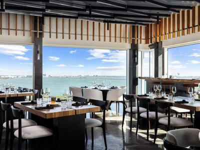 Modernes Restaurant mit Glasfronten und Blick auf das Meer und eine Stadt am Horizont.