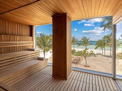 Sauna mit Holzbänken und Blick auf Strand und Palmen durch große Fenster.