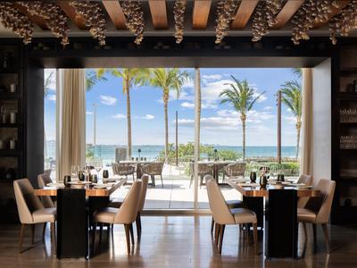 Modernes Restaurant mit großen Fenstern, Blick auf Pool und Palmen bei klarem Himmel.