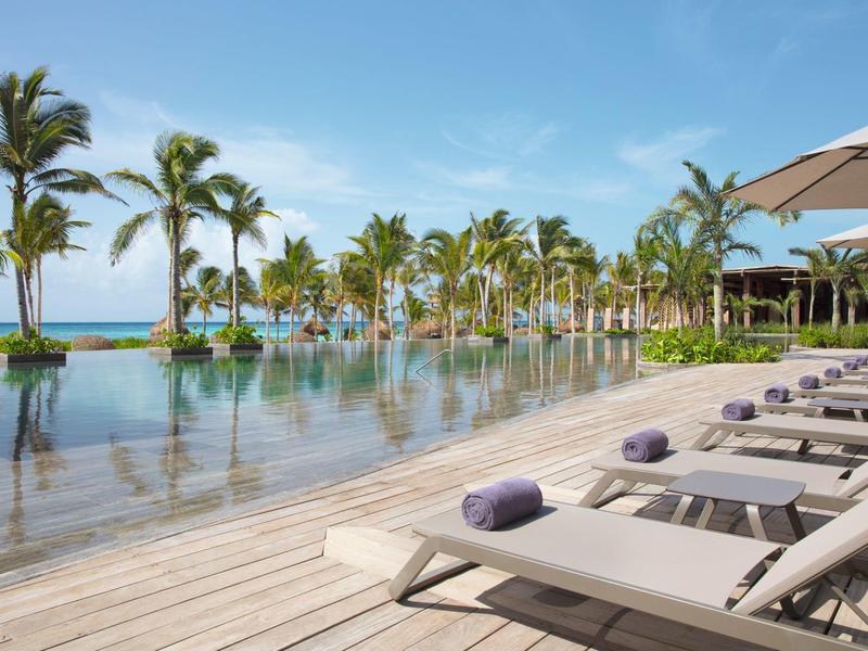 Infinity-Pool mit Liegestühlen, Handtüchern, Palmen und blauem Himmel am Meerufer.