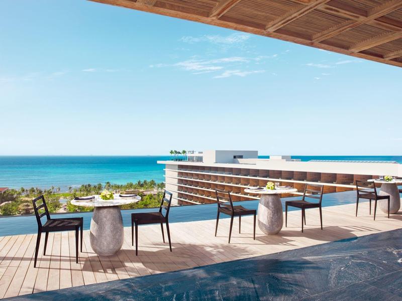 Terrasse mit Tischen und Stühlen, Blick auf Ozean, klarer Himmel, modernes Design, Holzdach.