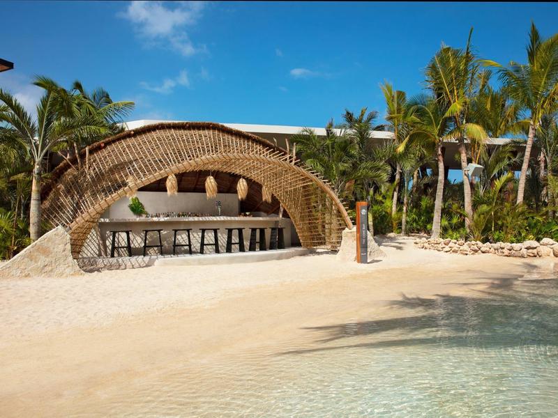 Strandbar mit gebogenem Dach aus Holz und Palmen im Hintergrund, Sand und klares Wasser im Vordergrund.