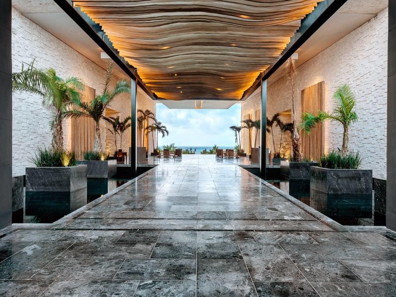 Langer heller Flur mit Marmorfußboden, Holzdecke, großen Pflanzen und Blick auf Meer und Himmel.