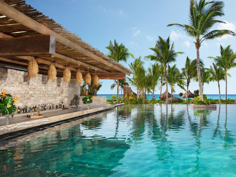 Schwimmbad mit türkisfarbenem Wasser, Palmen, Steinhütte und klarem Himmel am Meer.