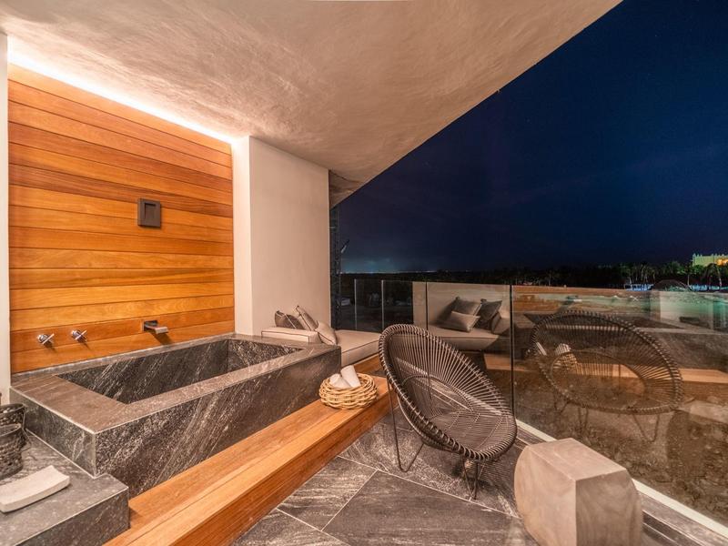 Moderner Balkon mit grauem Steinboden, Holzwand, freistehender Badewanne und Stühlen bei Nacht.