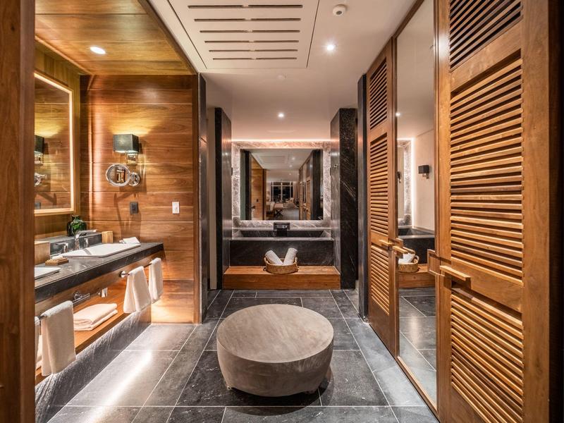 Schmaler Luxusbadezimmer mit dunklem Steinboden, Holzverkleidung, großem Spiegel und rundem Sitzhocker.