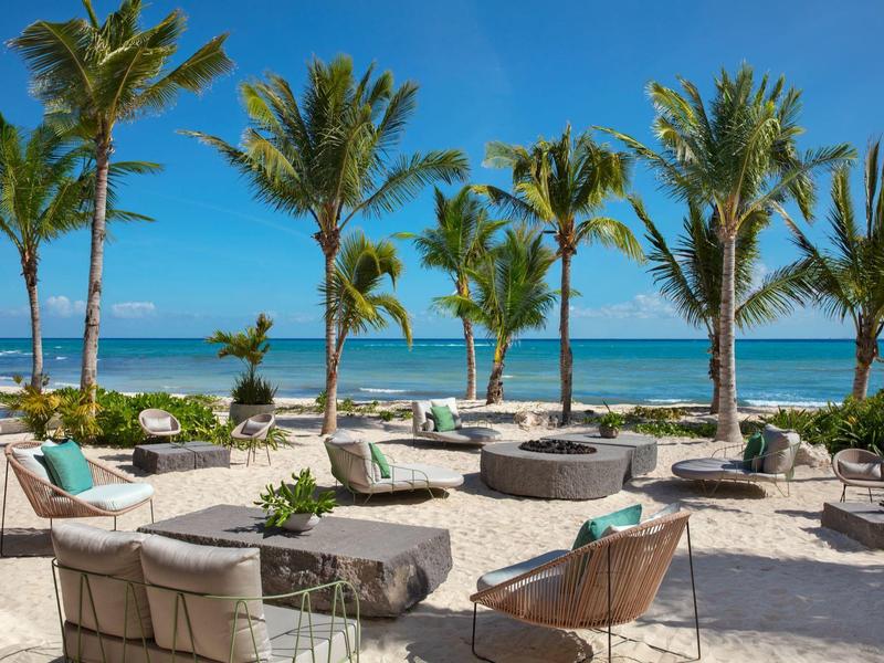 Strandterrasse mit Sitzgruppen, Palmen, Sandboden und blauem Meer im Hintergrund.