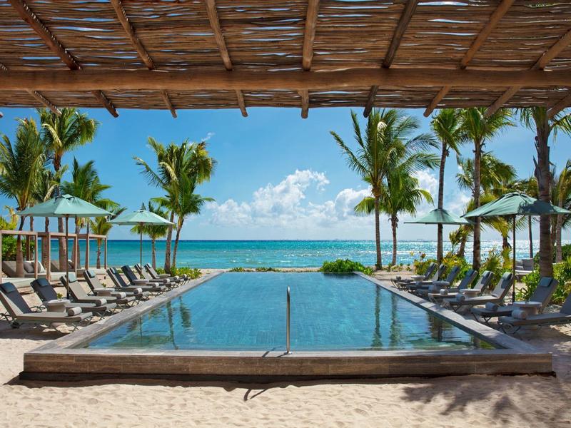 Pool mit Liegestühlen am Sandstrand, Palmen und klarem blauem Himmel im Hintergrund.
