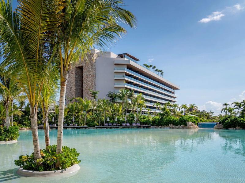 Modernes Hotel mit weißen Balkonen, Palmen und klarem blauem Wasser im Vordergrund.