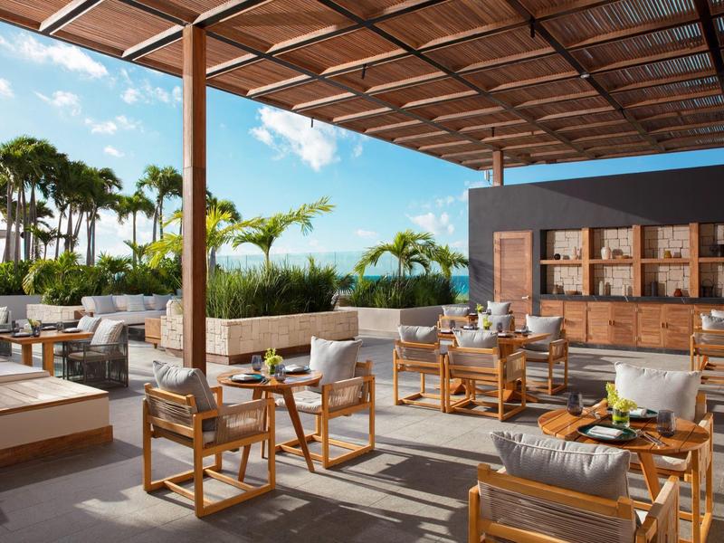 Moderne Außenlounge mit Holzstühlen, Sofas, grünen Pflanzen und Blick auf blauen Himmel.