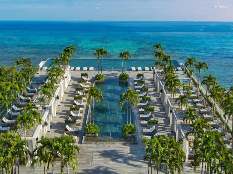 Blick von oben auf Palmen, ein rechteckiges Wasserbecken und Reihen moderner Bungalows am Meer.